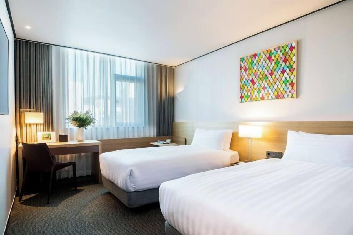 Une chambre d’hôtel avec deux lits, un bureau, une chaise, une lampe et une œuvre d’art colorée au mur.