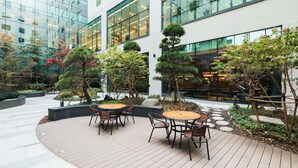 A modern outdoor seating area with round tables and chairs, surrounded by landscaped gardens and trees.