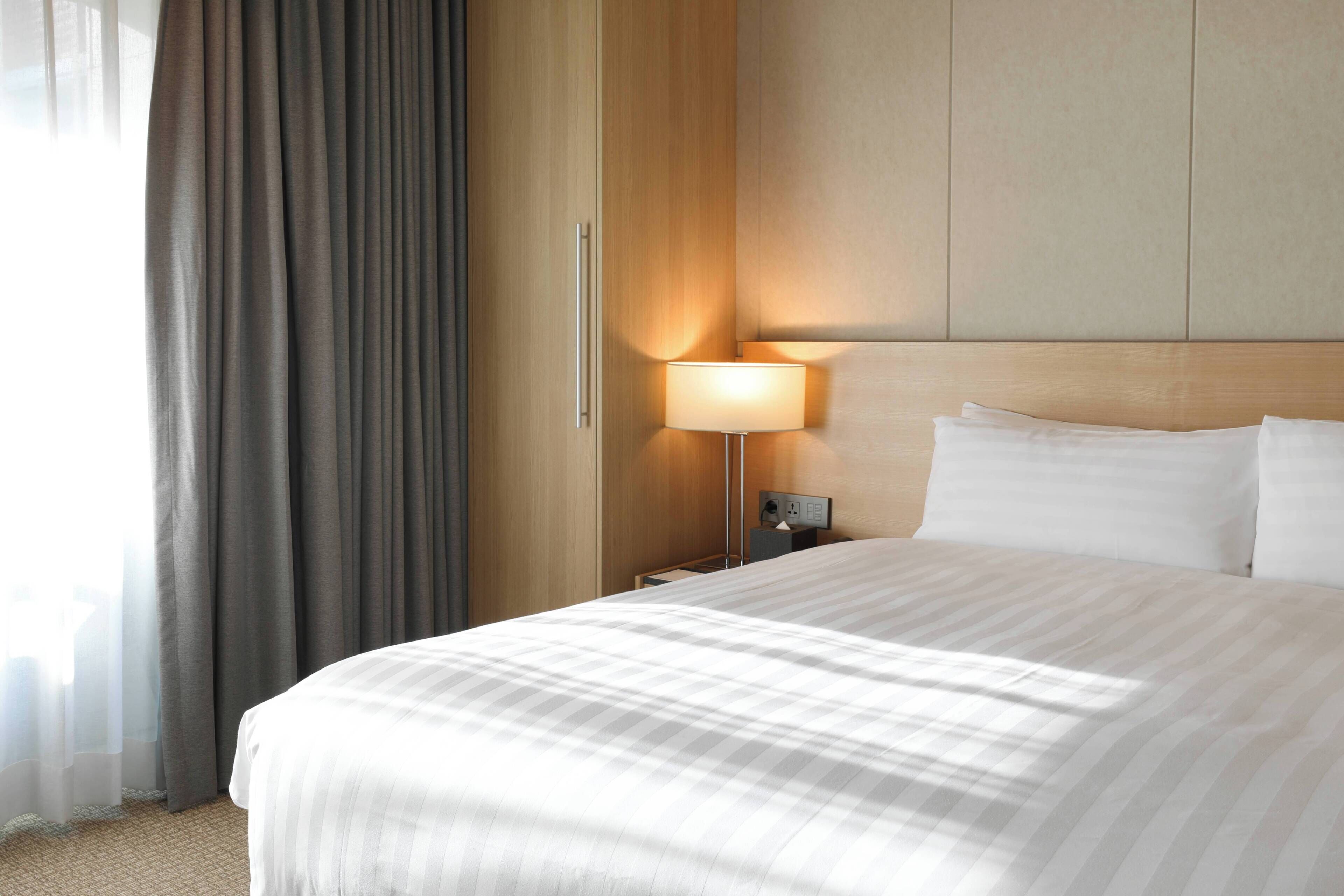 Une chambre d’hôtel avec un grand lit, une table de chevet avec une lampe, une tête de lit en bois et une vue sur l’extérieur à travers des rideaux transparents.