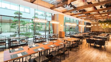 A spacious restaurant with high ceilings, exposed beams, and large windows offering views of greenery outside. Tables are neatly arranged with place settings.