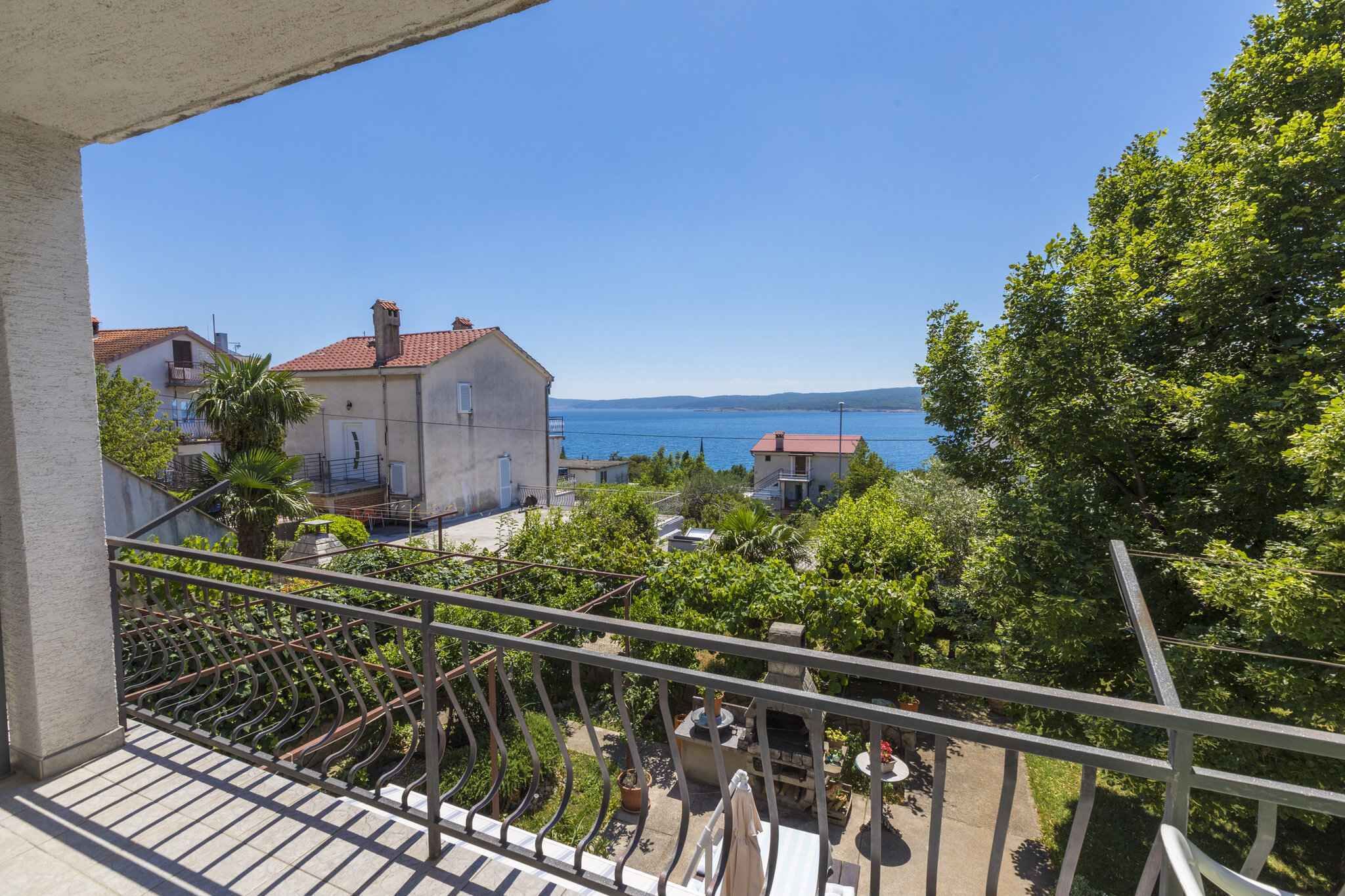 A balcony with a view of the sea, a garden, and a building with a red roof.