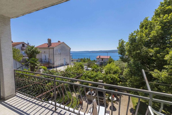 A balcony with a view of the sea, a garden, and a building with a red roof.