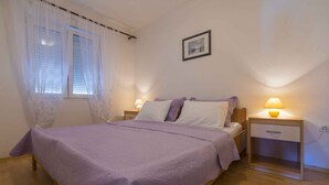 A bedroom with a bed, two bedside tables with lamps, a window with sheer curtains, and a framed picture on the wall.