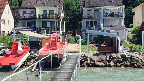 Una propiedad frente al agua con un muelle de madera que conduce a un bote rojo, rodeada de casas de varios pisos con balcones y áreas de estar al aire libre.