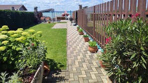 Un sendero en un jardín con una cerca de madera, plantas en macetas y una vista de un edificio al fondo.