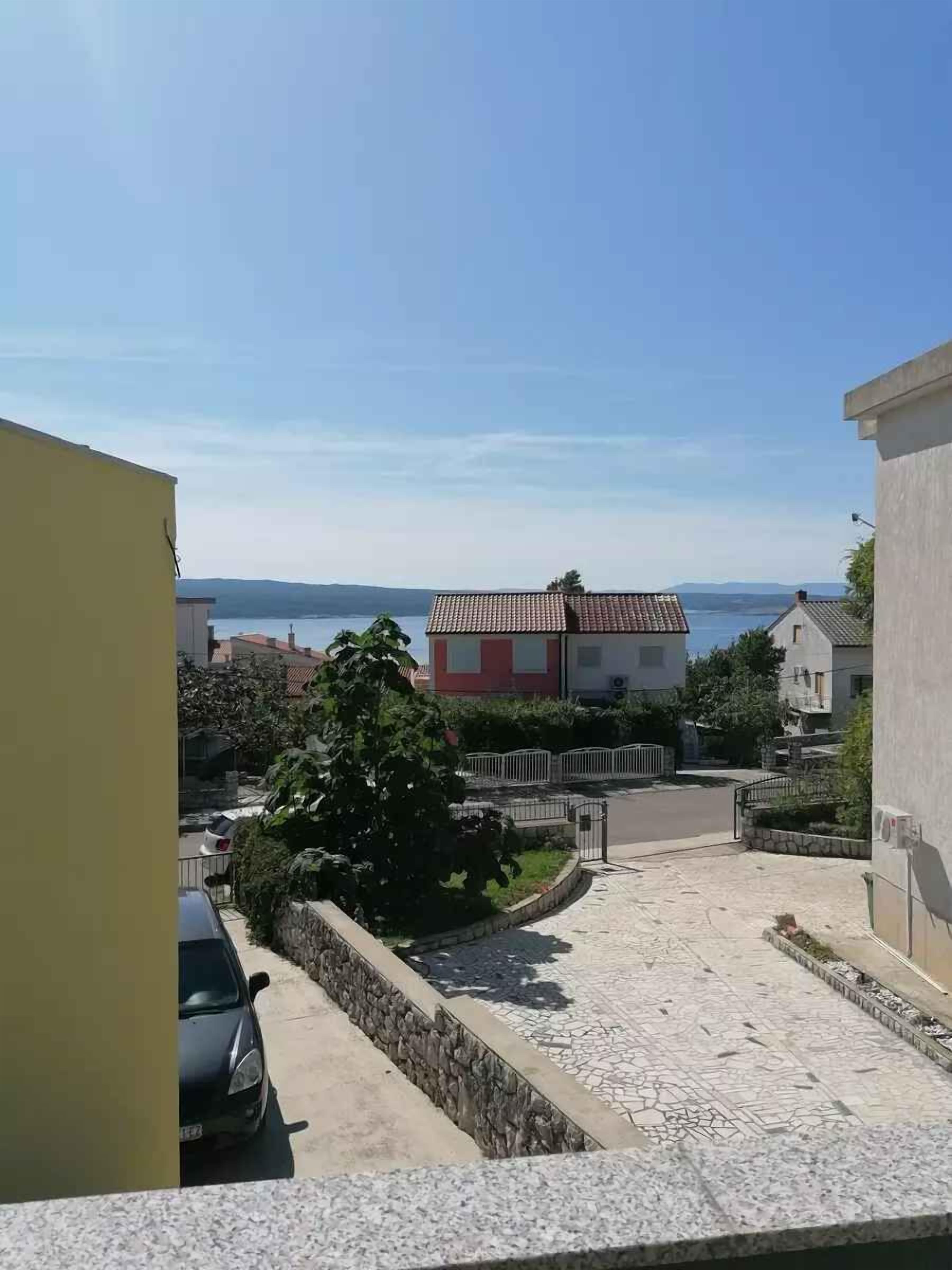 A residential area with a paved driveway leading to a house with a red and white facade, surrounded by greenery and a clear view of the sea in the background.