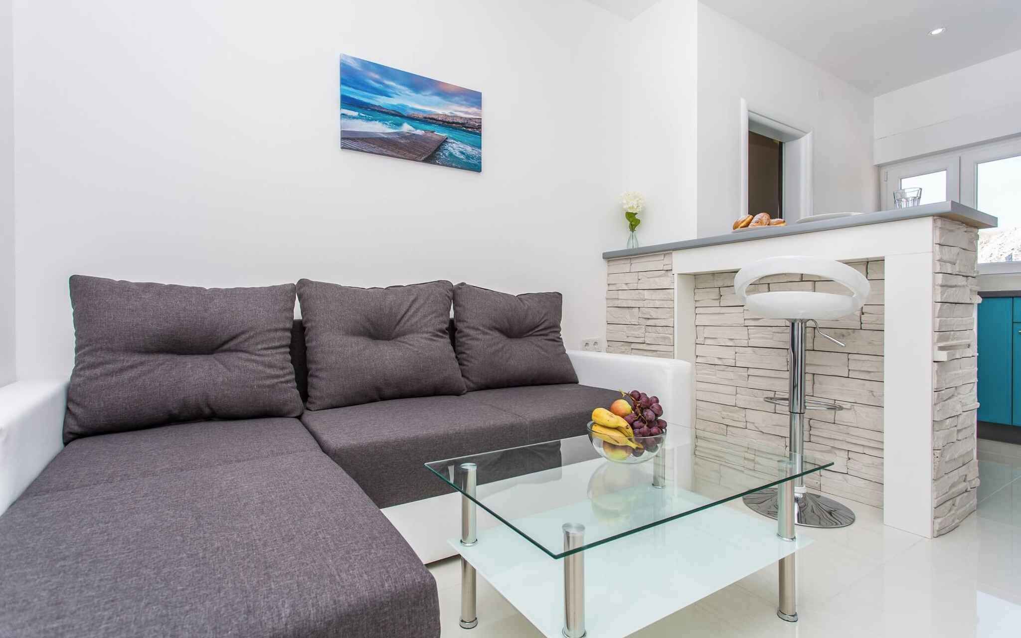 Ein modernes Wohnzimmer mit einem grauen Modulsofa, einem Glastisch mit Obst und einer Küchenzeile mit Steinrückwand und weißen Geräten.