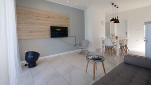 A modern living room with a flat-screen TV mounted on a wooden panel, a dining area with a white table and chairs, and a grey sofa.