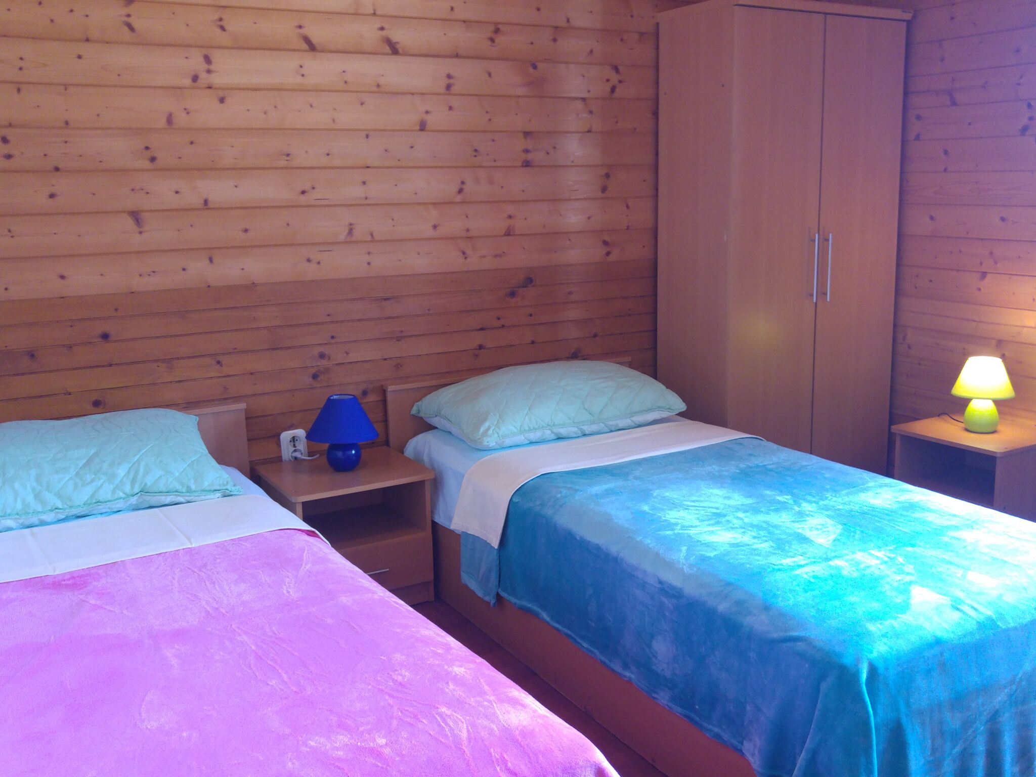 Quarto estilo cabana com duas camas, paredes de madeira, guarda-roupa e mesa de cabeceira com abajur.