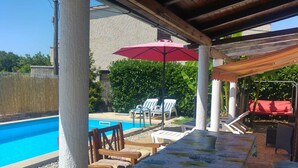 A covered outdoor area with a pool, wooden chairs, a table with a floral tablecloth, and a red umbrella. - Holiday home with washing machine and smart TV (Valbandon)