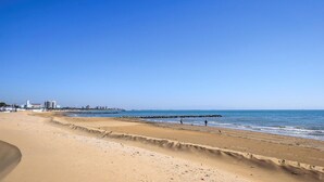 Een breed zandstrand met een helderblauwe lucht, een rotsenrij en gebouwen in de verte.