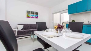 Un comedor moderno con una mesa blanca, sillas negras y un sofá gris. Hay tres cuadros en la pared, una ventana con vistas, y armarios de cocina color turquesa.
