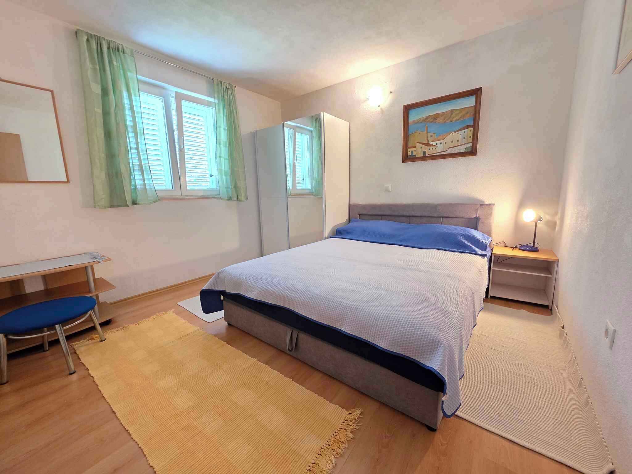 Un dormitorio con cama, escritorio, silla, mesita de noche con lámpara y un cuadro en la pared.
