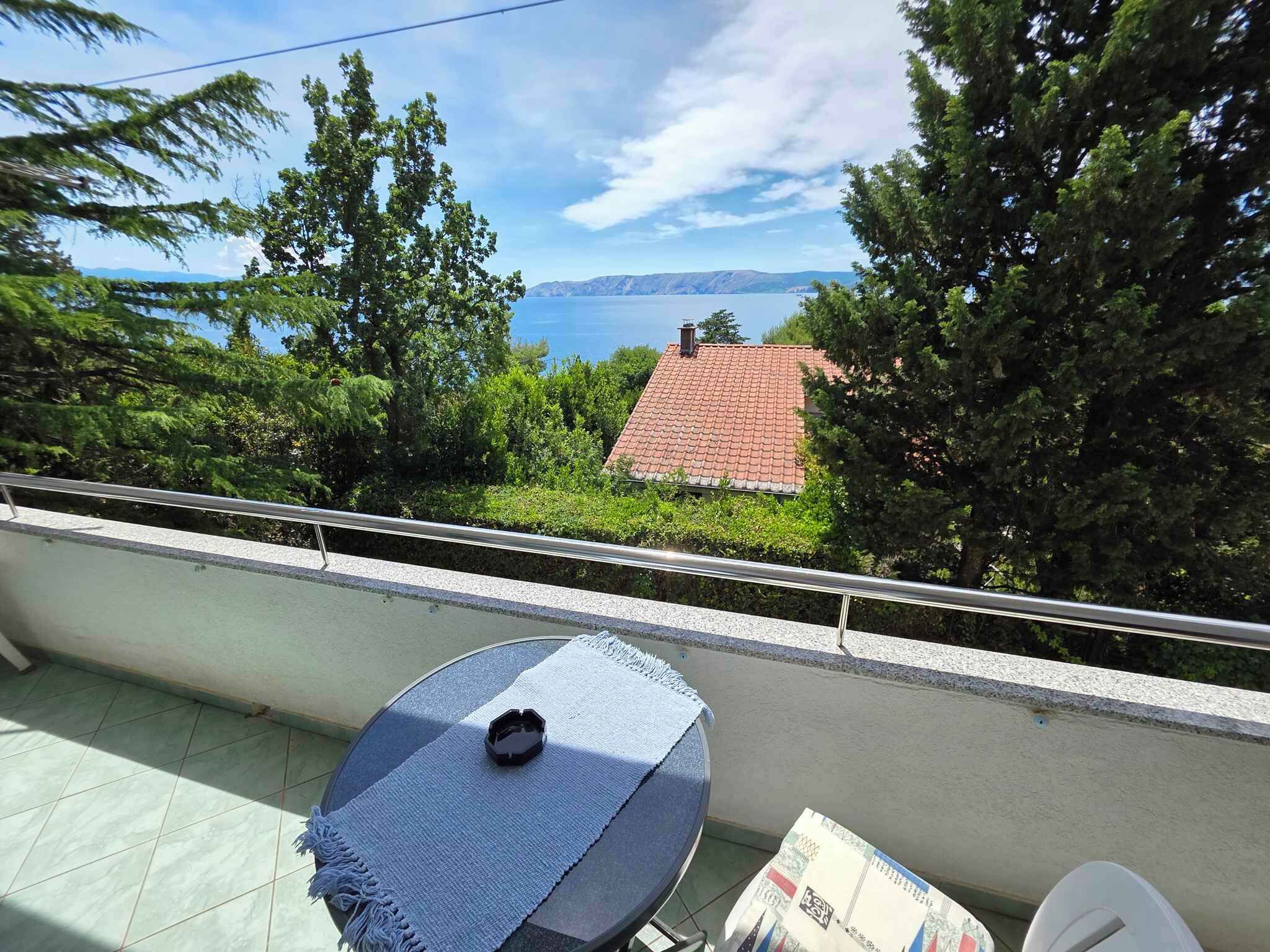A balcony with a table and chairs, overlooking a house with a red roof and a body of water in the distance.