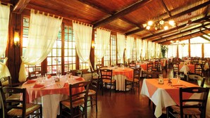 Een restaurant met meerdere tafels, houten balken en grote ramen met gordijnen.