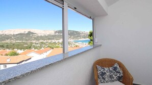 En balkon med kystlandskab i klart vue, en fletstol med hynde, og et vindue med persienner.