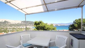 En balkon med bord og stole, udsigt over et smukt kystlandskab med bjerge og en klar himmel.