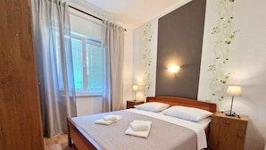 A neatly made bed with two pillows and folded towels, a bedside table with a lamp, and a window with curtains. - Holiday apartment with terrace and air conditioning (Baška)
