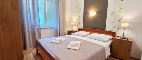 A neatly made bed with two pillows and folded towels, a bedside table with a lamp, and a window with curtains.