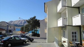 A building with a sign reading "AKZARIL" on the ground floor, a parking area with cars, and a view of mountains in the background. - Holiday apartment with terrace and air conditioning (Baška)