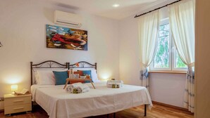 A neatly made bed with a white bedspread, pillows, and a tray with books and a cup. There is a nightstand with a lamp and a book, and a painting on the wall. - Villa with air conditioning (Campanedda/Sassari)