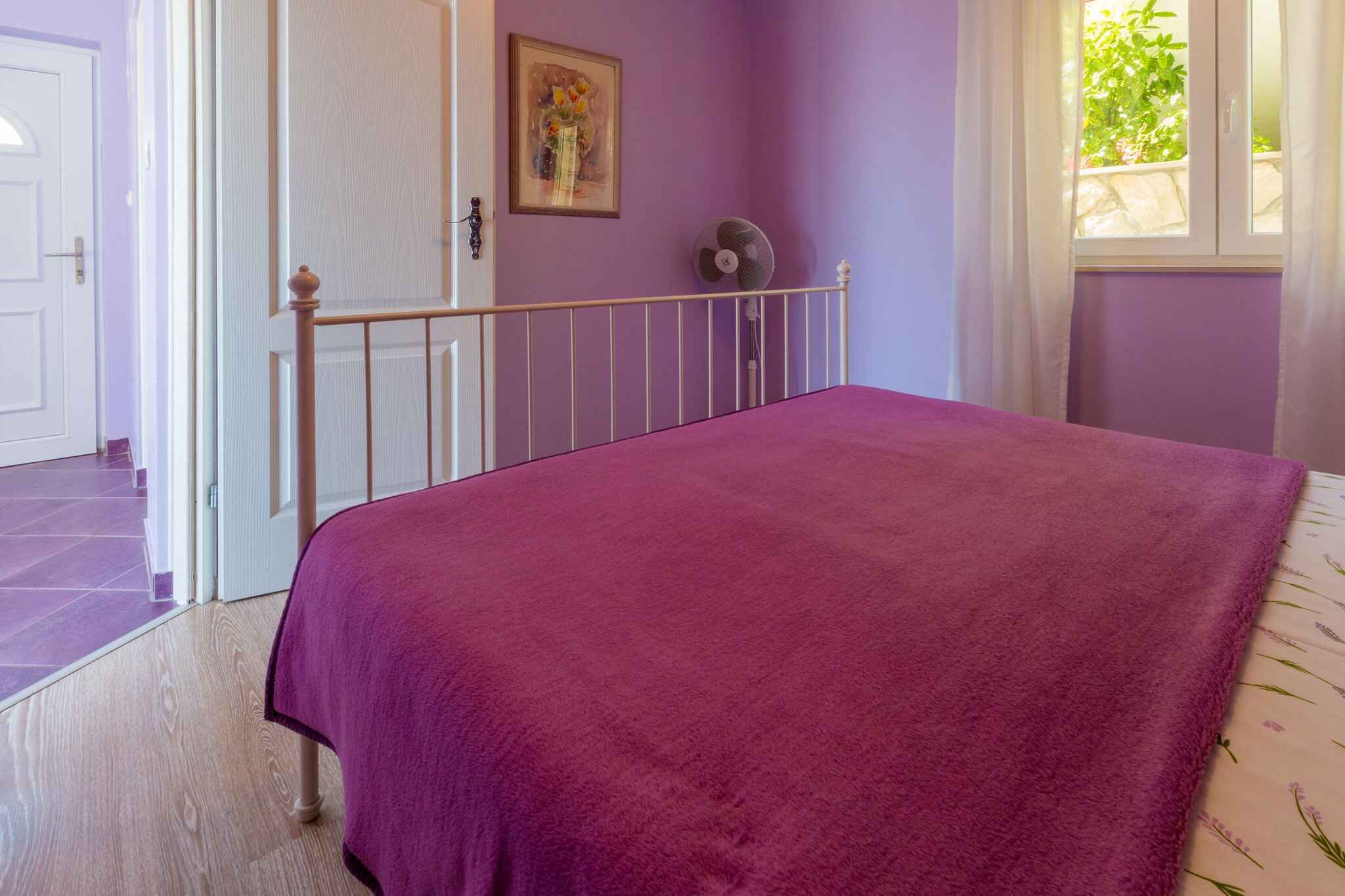 Ein Schlafzimmer mit violettem Überwurf, einer weißen Tür, einem Ventilator und einem Fenster mit Vorhängen.