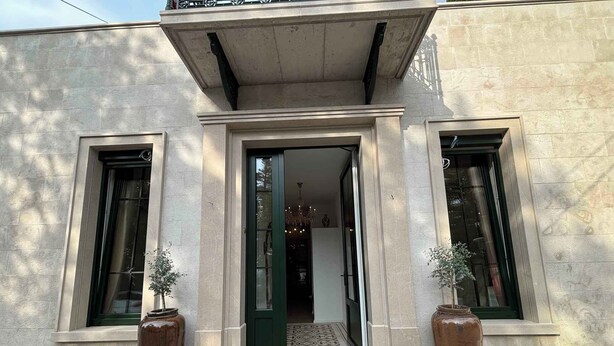 Entrada de um edifício com porta verde, duas janelas grandes e dois vasos de plantas.