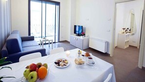 A modern living room with a sofa, a dining table with fruit and pastries, a TV, and a white cabinet.