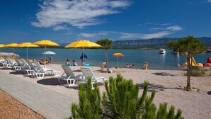 Ein Strand mit Liegestühlen und Sonnenschirmen, ein Holzsteg und der Blick auf das Meer und die Berge.