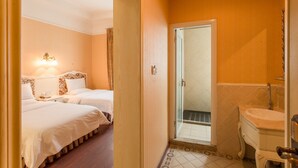 A hotel room with two beds, a bathroom with a sink and mirror, and a door leading to a shower area.