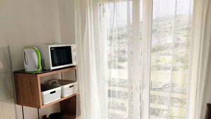 Una habitación con un estante de madera que contiene un microondas y un hervidor, un cubo de basura blanco y una ventana con cortina translúcida.