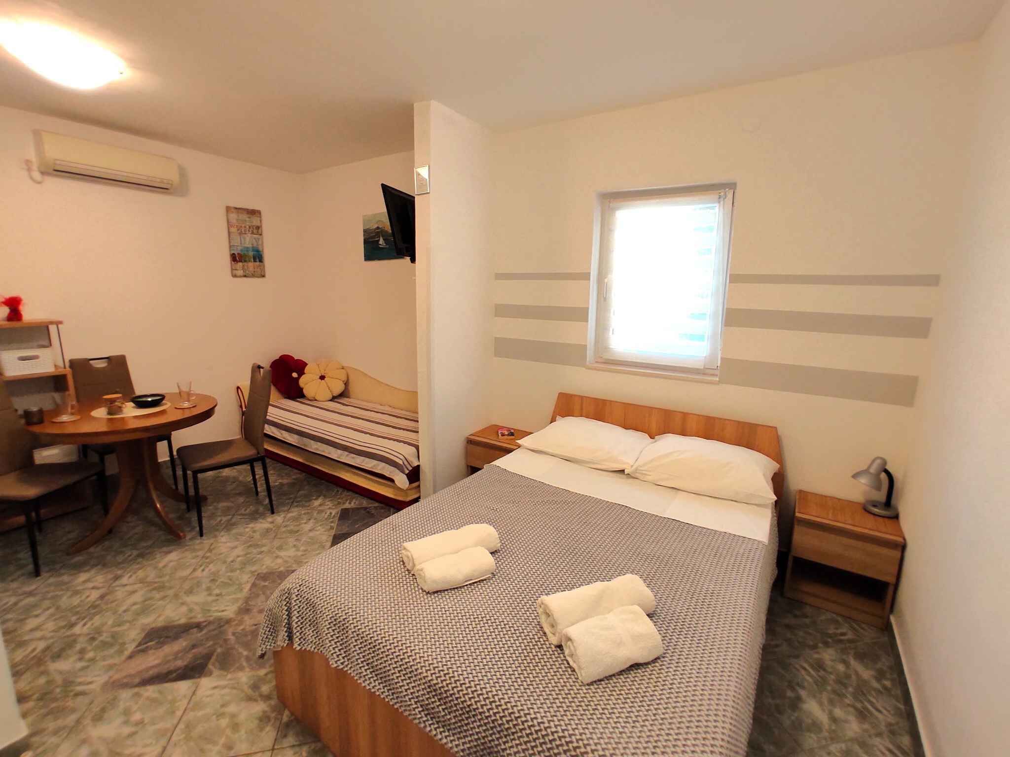 Ein kleines Hotelzimmer mit Bett, Nachttisch, einem kleinen Schreibtisch mit Stuhl, einem Fenster und einer Klimaanlage.