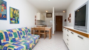 Un ambiente compatto con un divano colorato, un'area pranzo con sedie in legno, una cucina con mobili bianchi e un televisore sul mobile.
