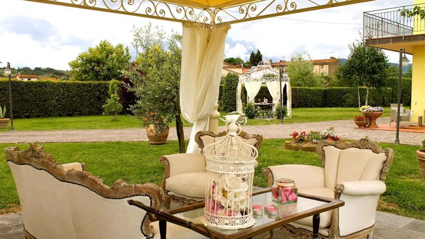 Een overdekt terras met twee stoelen en een tafel, een decoratieve vogelkooi, en uitzicht op een tuin.