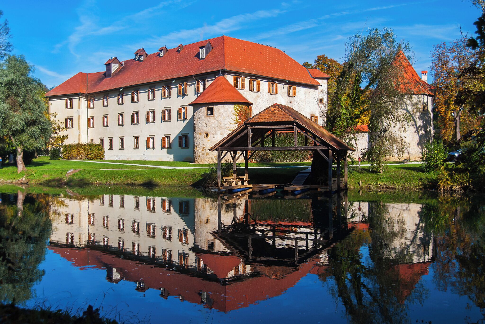 A large building with a red roof, a pond in front, and a wooden structure on the pond.