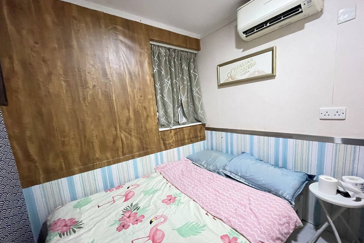 A bedroom with a bed, a window with curtains, an air conditioner, a wall-mounted sign, and a small table with a roll of toilet paper.