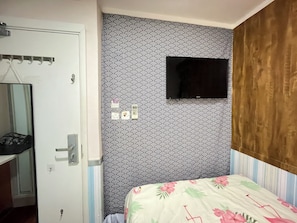 Et soveværelse med seng, ophængt tv, dør med knagerække og tapet.