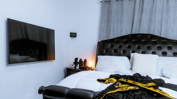 Een netjes opgemaakt bed met een zwart hoofdbord, een nachtkastje met een lamp en een aan de muur bevestigde televisie.