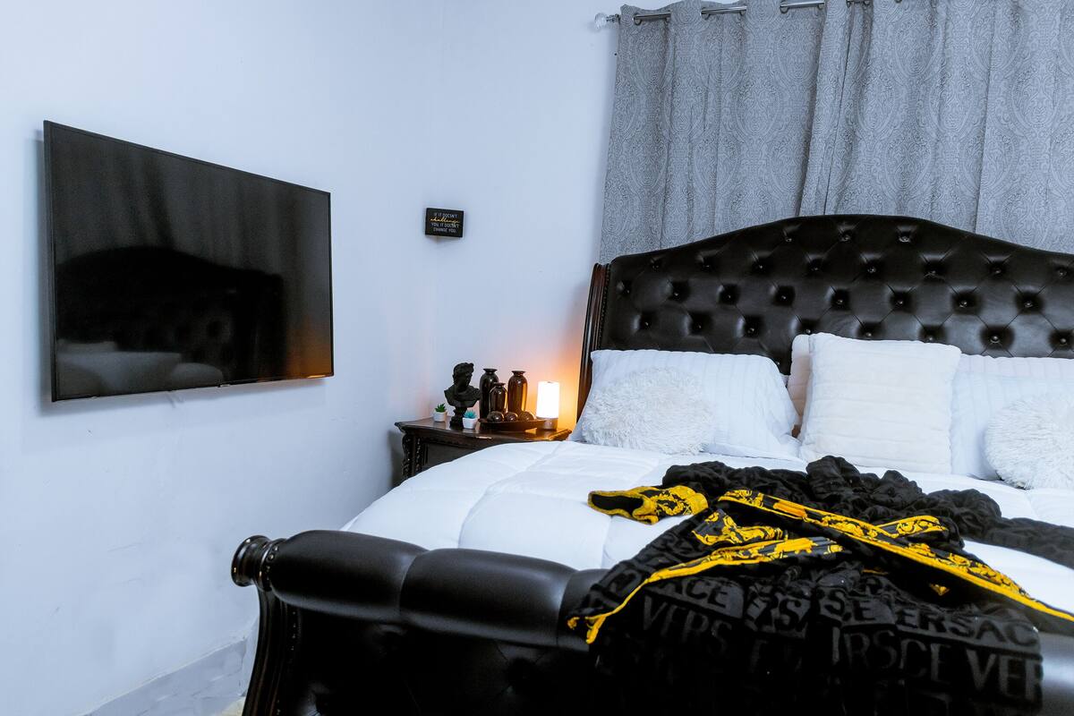 Un lit bien fait, avec une tête de lit noire, une table de chevet avec une lampe et un téléviseur fixé au mur.