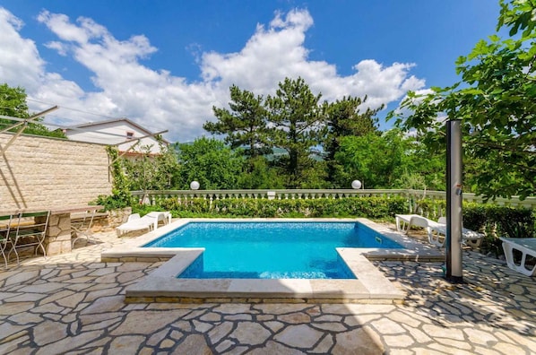 Une piscine aux bords recouverts de pierres, entourée de verdure et agrémentée de chaises longues blanches.