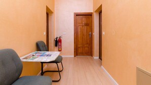 A narrow hallway with a wooden door at the end, a fire extinguisher mounted on the wall, and a table with magazines in the foreground.