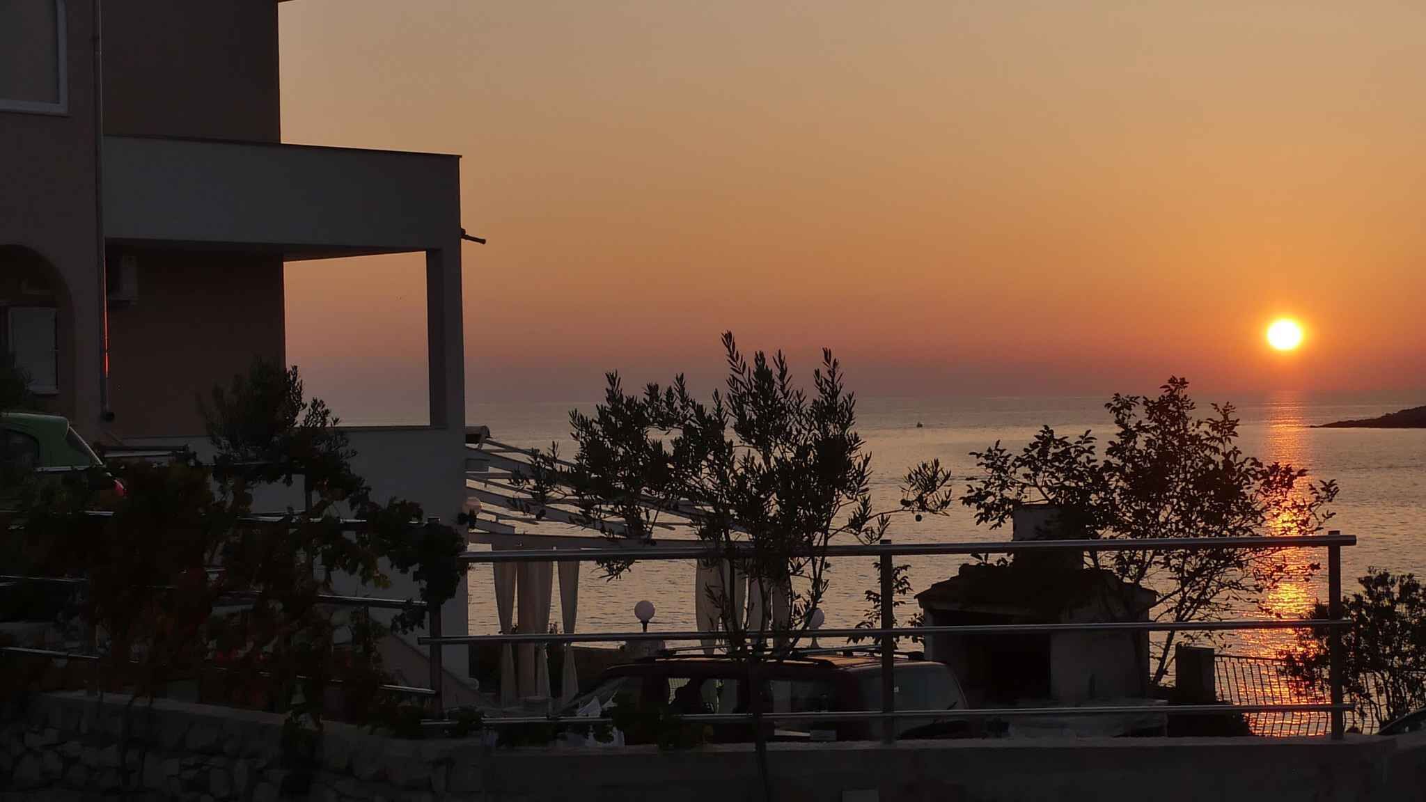 A coastal view with a sunset, a building with a balcony, and a parked car.