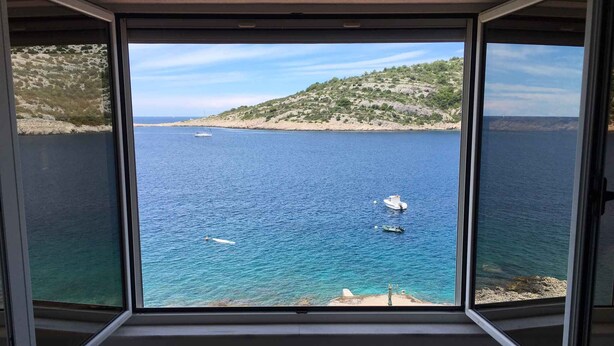 Vista desde una habitación con ventanas amplias que dan a un mar tranquilo, con barcos, una playa de arena y un paisaje montañoso.