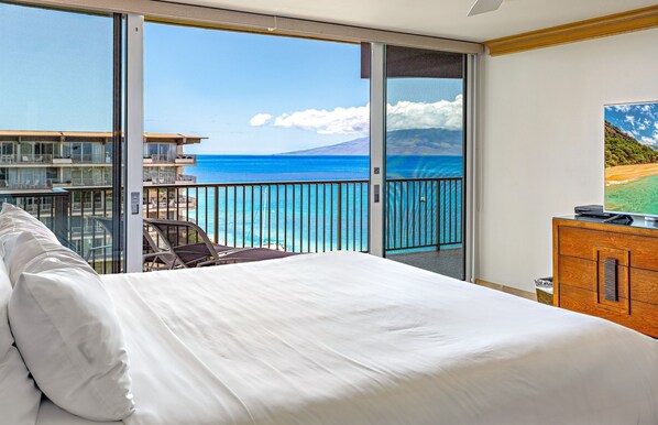 Een hotelkamer met een groot bed, een televisie en een balkon met uitzicht op de oceaan en een berg.