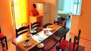 Un comedor con una mesa dispuesta para dos, un refrigerador, una cama al fondo y una ventana con cortinas.