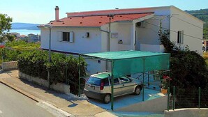 Una casa de dos pisos con un coche aparcado bajo un toldo verde, techo de tejas rojas y vista al mar en el fondo.