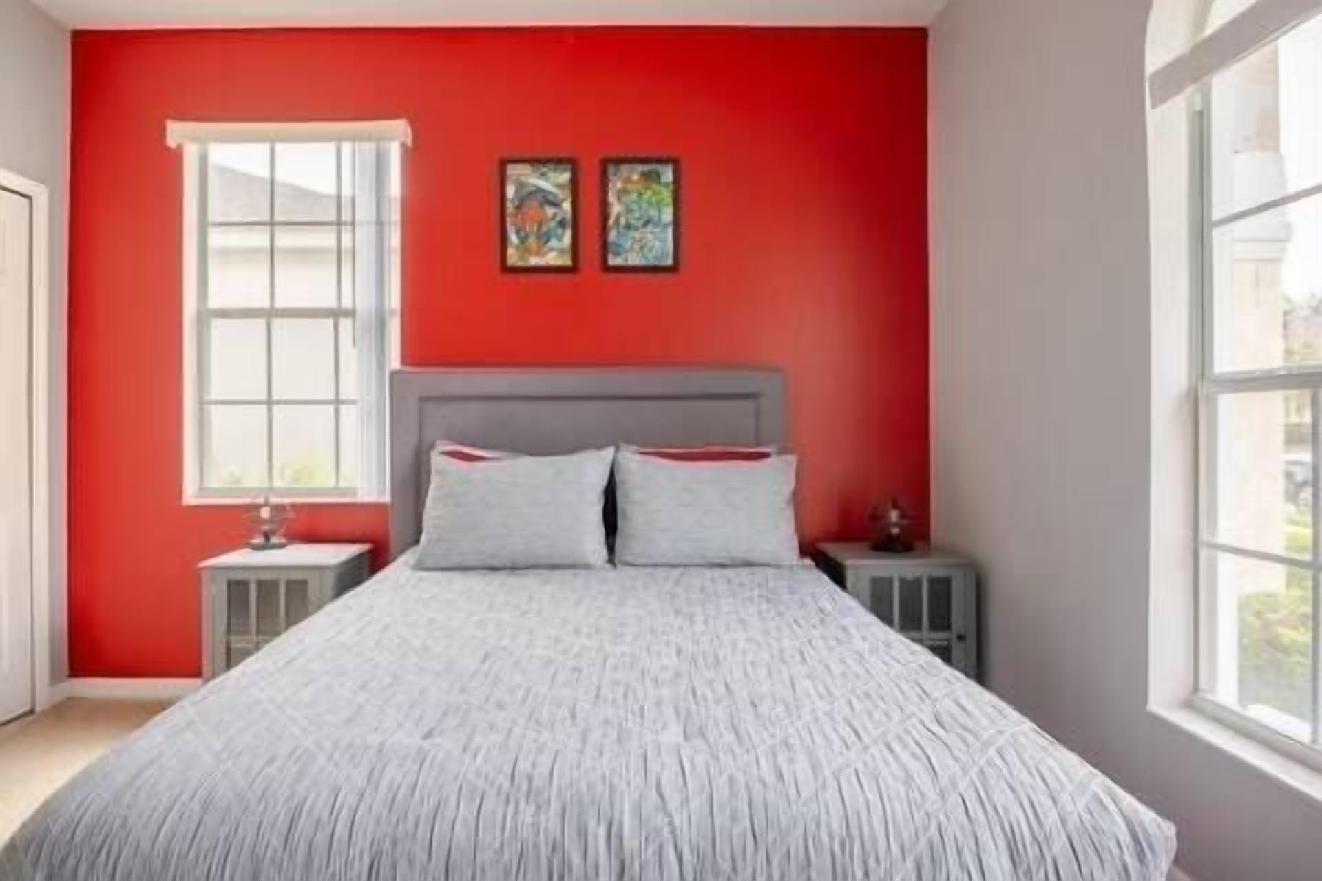 A bedroom with a red wall, a grey bed, two pillows, a window, and two artworks on the wall.