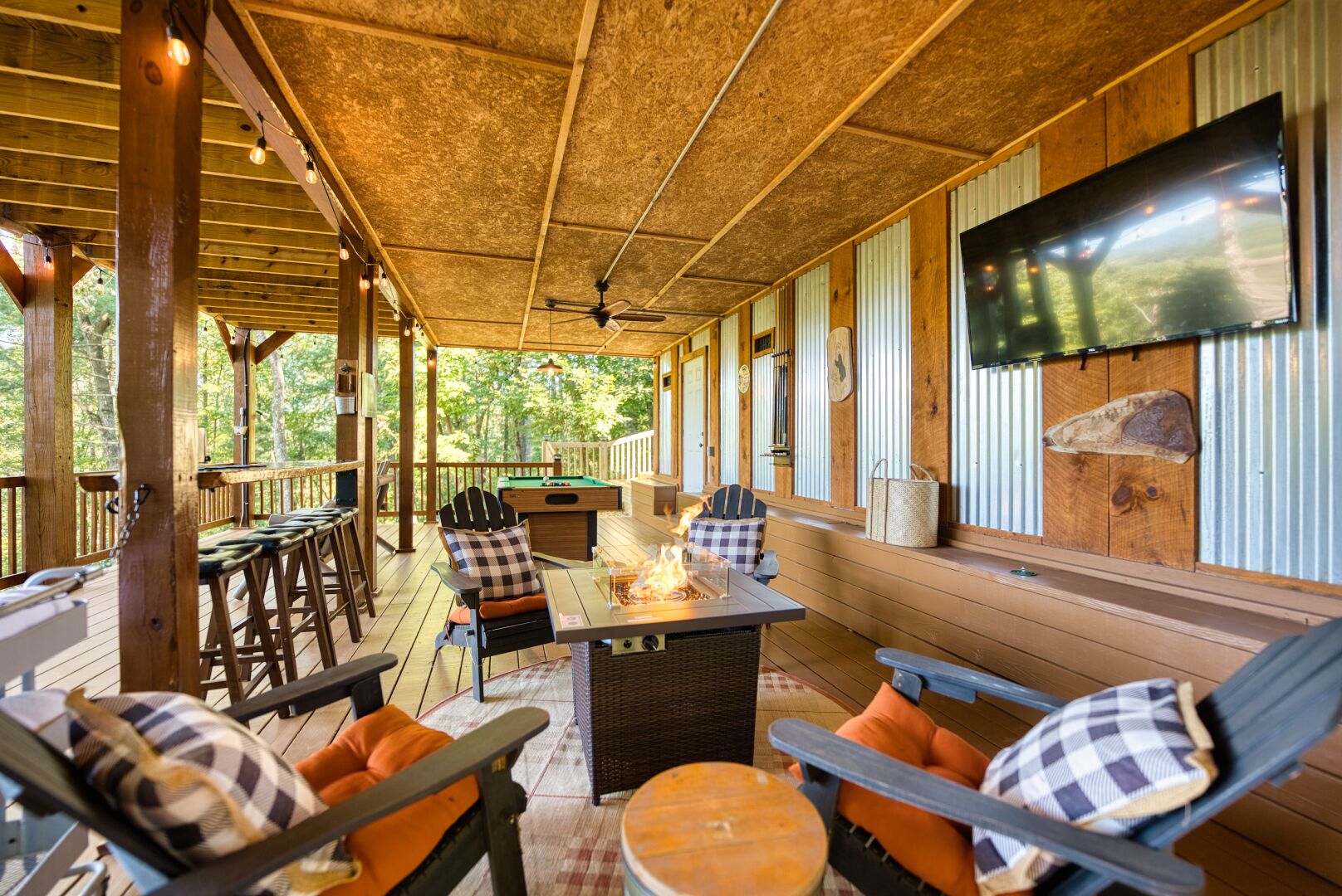 A covered outdoor deck with wooden furniture, a pool table, and a flat-screen TV mounted on the wall.