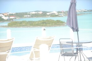 Zona de piscina con sillones de descanso y una mesa, con vistas al mar turquesa y edificios a lo lejos.