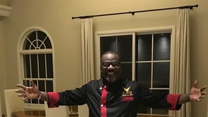 Um homem de camisa preta com mangas vermelhas está em pé num quarto com uma mesa de jantar e cadeiras, uma janela com cortinas e chão de azulejos.